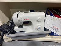 White Singer Tradition 2259 sewing machine placed in gray plastic tray with foot pedal and power cord visible beside it. Instruction manual for Singer Tradition 2259 visible in background.