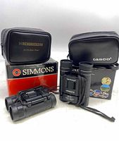 Two pairs of binoculars displayed with their respective black carrying cases and original packaging boxes. Tasco Voyager II binoculars in upright position with strap visible, and Simmons compact binoculars lying horizontally in front.