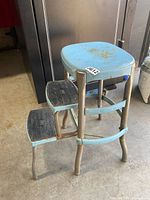 Side and front perspective of vintage robin egg blue Cosco folding step stool showing rusted metal frame, two black rubberized steps and curved legs.