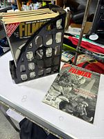 Side view of a stack of approximately 28 Filmax magazines in a black plastic magazine holder, displaying various covers and spine details, on a white table with surrounding tools and items in background.