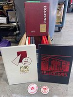 Photo showing the 1971 maroon yearbook, 1984 black yearbook, 1990 white yearbook, and two round white buttons with red graphics and text related to North Catholic High School Falcons.