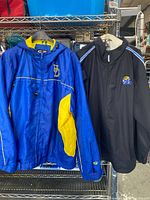 Two University of Delaware football jackets hanging on hangers, blue/yellow hooded windbreaker and black pullover with football helmet embroidery.