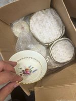 Hand holding a Royal Doulton teacup showing floral motif and gold trim. Box contains matching bowls with fabric covers.