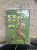 Front cover of 'The Babe Ruth Story' paperback showing Babe Ruth in a batting stance on a green background, inside a protective bag.