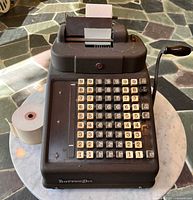 Front top view of Burroughs adding machine with installed paper, spare tape roll beside