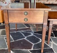 Front view of one end table showing drawer with brass pulls and fluted legs