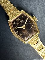 Close-up of the Bulova watch face showing the black dial, gold-tone Arabic numerals, hands, and textured gold-toned casing.