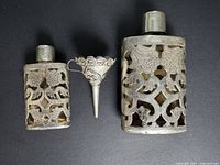 Three-piece set showing two carved sterling silver perfume flasks and an ornate silver funnel in the center, all laid on a black background.