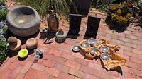 All items laid out on brick patio including stone basin, brass bell, Buddha plaques, bowls, wood rounds, frogs