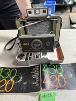 Front view of Polaroid 420 Automatic Land Camera open with bellows extended, strap visible, manuals in foreground