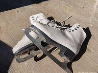 Pair of white figure skates on concrete surface showing blades and uppers