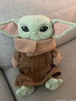 Front view of Baby Yoda plush toy sitting on couch, showing face, ears, brown robe with corduroy collar and cuffs, and feet.