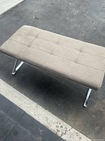 View of the full beige cushioned entrance bench on pavement, showing stitched pattern and chrome legs.