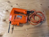 Orange Black & Decker electric jig saw laying on wooden surface, showing whole tool and power cord.