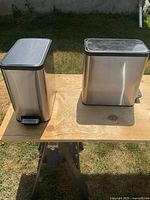 Two stainless steel garbage cans placed on a wooden board outdoors showing rectangular shape, black plastic lids and one with foot pedal.