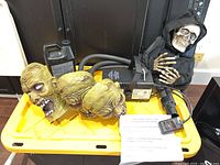 Fog machine unit with three zombie heads and bottle of fog juice displayed on yellow plastic lid, with animated crawling skeleton decoration in back.