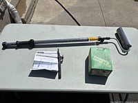 Wagner Paint Stick EZ-Twist roller laid flat with instruction manual and Mastercraft Sweeping Compound box beside it on a pale gray table outdoors.