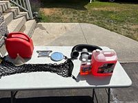 Photo showing all items on a white folding table outside, includes red gas can, funnel, black cargo netting, black hose, two liquid containers, shaving mirror, and wrenches.