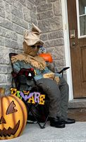 Full view of the sitting scarecrow seated on a folding chair on a porch with a 'Beware' sign and pumpkin decoration.