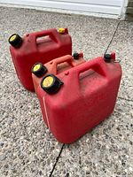 Two red gasoline containers standing upright on concrete surface, showing handles, caps, and molded lettering