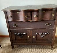 Front view of the full dresser showing the wavy top edge and drawer fronts, two panel doors with carved accents and two brass handles on the bottom drawer.