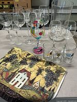 Overview of glassware set with large vase, wine glasses including one with colorful cartoon images, and dish towel with grapevine and house pattern.