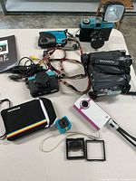 Overview of six cameras on table including Polaroid Cube with case, Diana F camera, Flip video camera, Magnavox camera, Canon camera, and one more compact camera