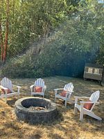 Four white Adirondack chairs arranged around a stone fire pit, each with a rust-colored throw pillow.