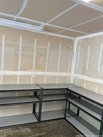 Three short metal garage shelving units placed in an L-shape in a garage with unfinished drywall, showing black metal frames and gray laminate shelves.