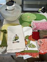 Image showing the plastic salad spinner, various kitchen towels including one embroidered with Slovenia and a gnome doll, alongside green leaf shaped rubber bowl covers.