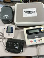 Photo of the weight scale with sign 'BARE FEET NOT PERMITTED ON SCALE.' with digital display panel and buttons, blood pressure monitor with attached cuff, and black hard carrying case.