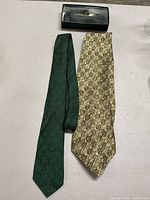 Two silk neckties and a boxed silk bowtie arranged side by side on a white surface. The dark green tie has a subtle diamond pattern, the lighter tie features a repeating animal motif, and the bowtie is in a black Pierre Balmain box.