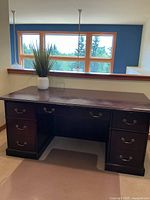Front view of the large wooden desk with brass handles and multiple drawers, showing the decorative plant on top and window background.