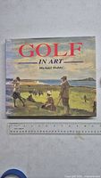 Front cover of the book titled Golf in Art by Michael Hobbs, showing an illustrated golf scene with players on a golf course and distant hills under a cloudy sky.