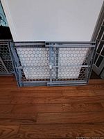 Two gray expandable plastic baby gates standing on hardwood floor, showing hexagonal mesh pattern and extendable sides.