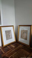 Two framed sketches with gilded wood frames and double mats leaning against a wall on a wooden floor. One sketch is female figure, the other male figure.
