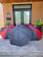 Three large golf umbrellas arranged on a porch step in front of a black double door, showing closed canopies with two red and black striped and one solid black umbrella.