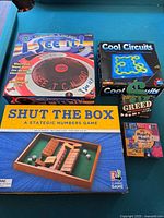 Five games shown on turquoise background: I See It, Cool Circuits, Shut The Box, Greed dice game, and Mighty Magic kit unopened.
