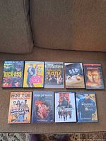 Top view of the front covers of 10 DVD movies arranged in two rows on a couch showing titles including Kick-Ass, Austin Powers, American Hustle, Bad Teacher, Enemy at the Gates, Hot Tub Time Machine, Across the Universe, Inglorious Basterds, The Bourne Identity.