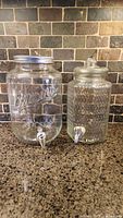 Photo of two clear glass drink dispensers with spigots on a kitchen counter, mason jar style on left with metal lid and bubbly textured one on right with glass lid.