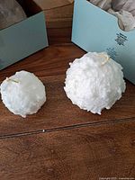 Two snowball candles, one 2-inch and one 3-inch, displayed side by side on a wooden surface with original packaging boxes in the background.