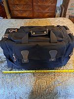 Front and top view of black Rothco large canvas travel bag showing size and main zipper compartment with two front buckle pockets, placed on bed with ruler for scale.