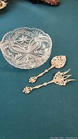 Photo showing full lot - clear cut crystal bowl with four feet and two ornate Victorian style metal serving utensils on green surface.