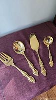 Top view of 4 gold plated serving utensils on purple cloth: serving fork, large serving spoon, pie server, shorter gravy ladle