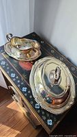 Gravy boat on a matching silver plated underplate and covered vegetable server with lid and detachable handle placed side by side on a painted cabinet.