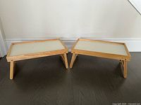 Two IKEA wooden lap tables with raised edges and foldable legs, placed on a dark hardwood floor against a plain white wall.