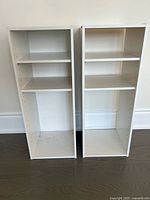 Two white IKEA shelves side by side against a wall on a wooden floor, showing front view with visible adjustable peg holes and some wear marks.