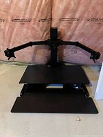 Front view of black sit/stand desk converter with triple monitor arm mount on top, black surface platforms visible, motor mechanism under desk surface.