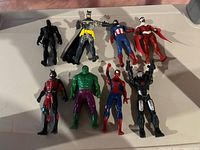 Eight superhero action figures arranged on a beige surface including Black Panther, Batman, Captain America, Spider-Man, Hulk, Ant-Man, and two others.