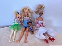 Four dolls arranged sitting: one Tinkerbell fairy doll, two Barbie dolls, and one baby Barbie doll.